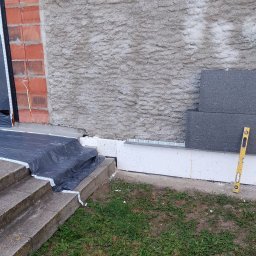 P&K BUD Ewa Kotlińska - Fragment elewacji w trakcie ocieplania styropianem, widoczne płyty izolacyjne i poziomica. Część ściany pokryta szarym tynkiem, zabezpieczone schody.