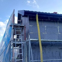 P&K BUD Ewa Kotlińska - Elewacja domu w trakcie ocieplania, widoczne rusztowanie i żółty przewód wentylacyjny, ściana pokryta ciemnoszarymi płytami izolacyjnymi.