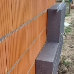 Fragment ściany z pustaków ceramicznych i szarego ocieplenia styropianowego, widoczne detale łączenia materiałów, grunt z roślinnością w tle.