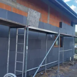 Ocieplanie budynku styropianem grafitowym, widoczne rusztowanie i fragment ściany z cegły. Częściowo ocieplona elewacja domu jednorodzinnego w trakcie prac.