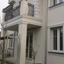 Elegancka elewacja domu z balkonem i ozdobną balustradą, widoczne detale sztukaterii i okien. Na pierwszym planie rusztowanie sugerujące prace wykończeniowe.