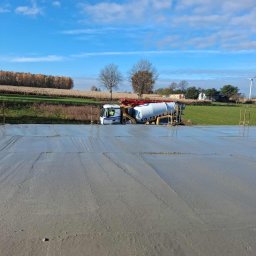 P&K BUD Ewa Kotlińska - Betoniarka wylewa beton na przygotowany teren budowy, widoczne zbrojenia i elementy konstrukcyjne. W tle krajobraz wiejski i błękitne niebo.