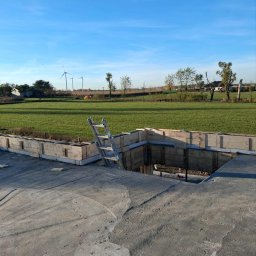 P&K BUD Ewa Kotlińska - Fundament domu w trakcie budowy: betonowa płyta, drewniane szalunki, drabina. W tle zielone pole i turbiny wiatrowe pod błękitnym niebem.