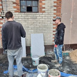 P&K BUD Ewa Kotlińska - Dwóch mężczyzn przy pracy na zewnątrz budynku z cegły i tynku. Widoczne narzędzia, wiadra z zaprawą i płyty izolacyjne. Prace elewacyjne w toku.