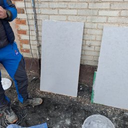 P&K BUD Ewa Kotlińska - Przygotowanie próbek elewacji w Kutnie: Dwie płyty z szarym tynkiem strukturalnym, wiadro z masą tynkarską i fragment muru z cegły w tle.