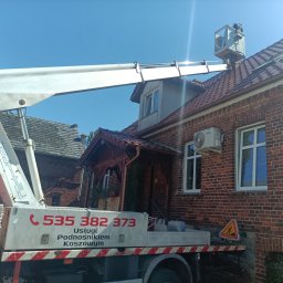 Przedsiębiorstwo handlowo usługowe Mariola Dyba - Pracownik w koszu podnośnika koszowego na dachu ceglanego domu podczas inspekcji. Widoczny fragment podnośnika z numerem telefonu i znak ostrzegawczy.