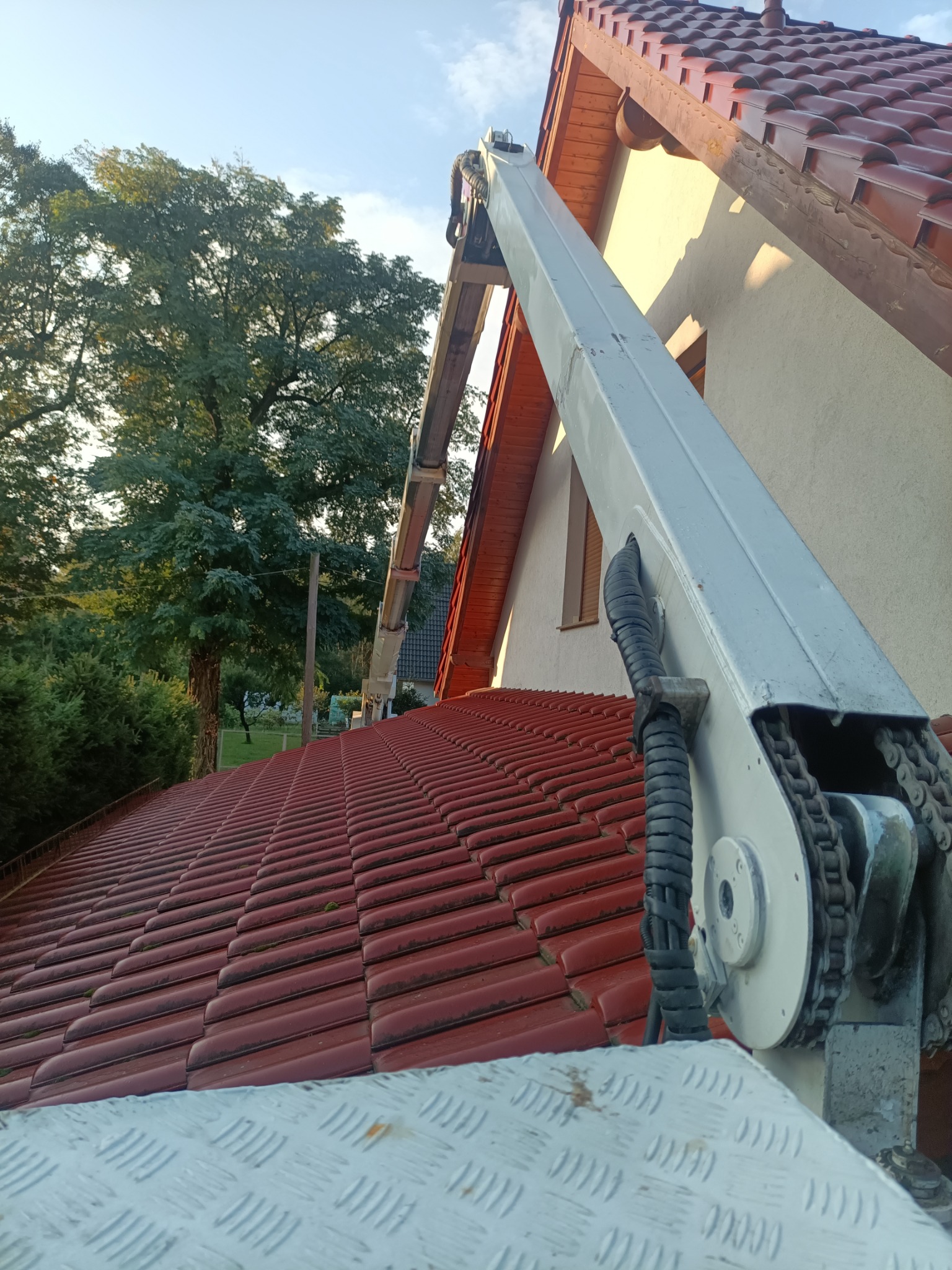 Podnośnik koszowy na tle dachu z czerwonej dachówki i zieleni drzew. Widok na prace wysokościowe z bliska, z perspektywy platformy roboczej.