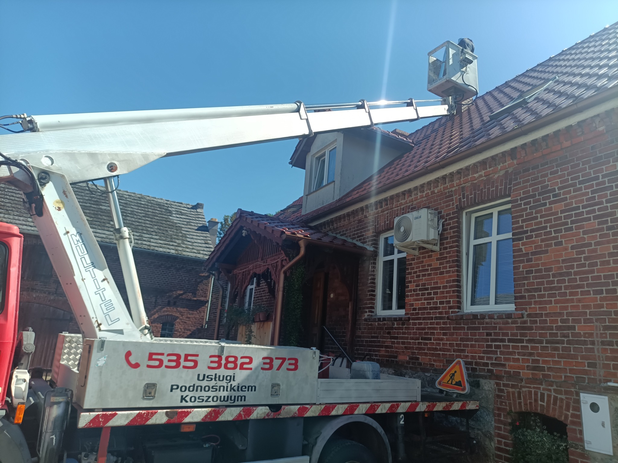 Pracownik w koszu podnośnika koszowego na dachu ceglanego domu podczas inspekcji. Widoczny fragment podnośnika z numerem telefonu i znak ostrzegawczy.