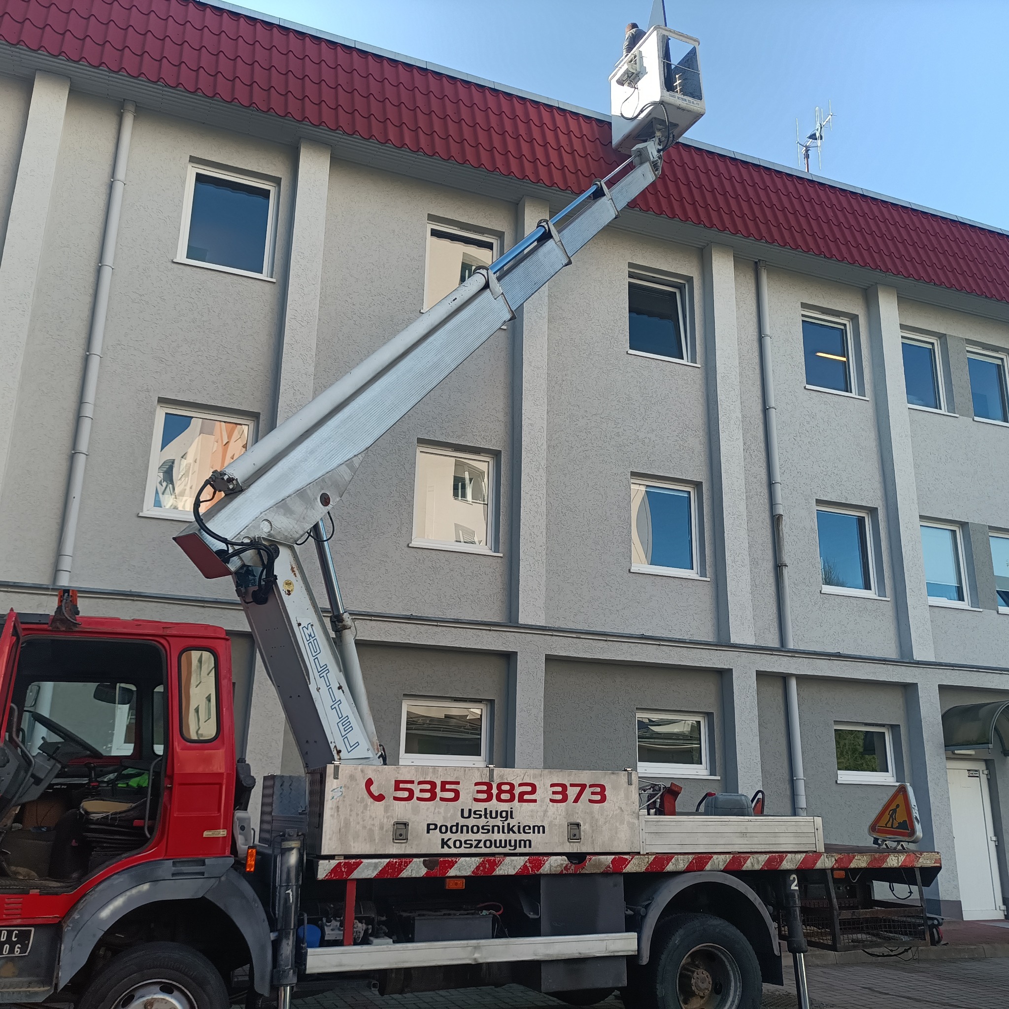 Czerwony podnośnik koszowy przed szarym budynkiem z czerwoną dachówką, antena na dachu. Na koszu osoba w oddali, widoczne numery telefonu na pojeździe.