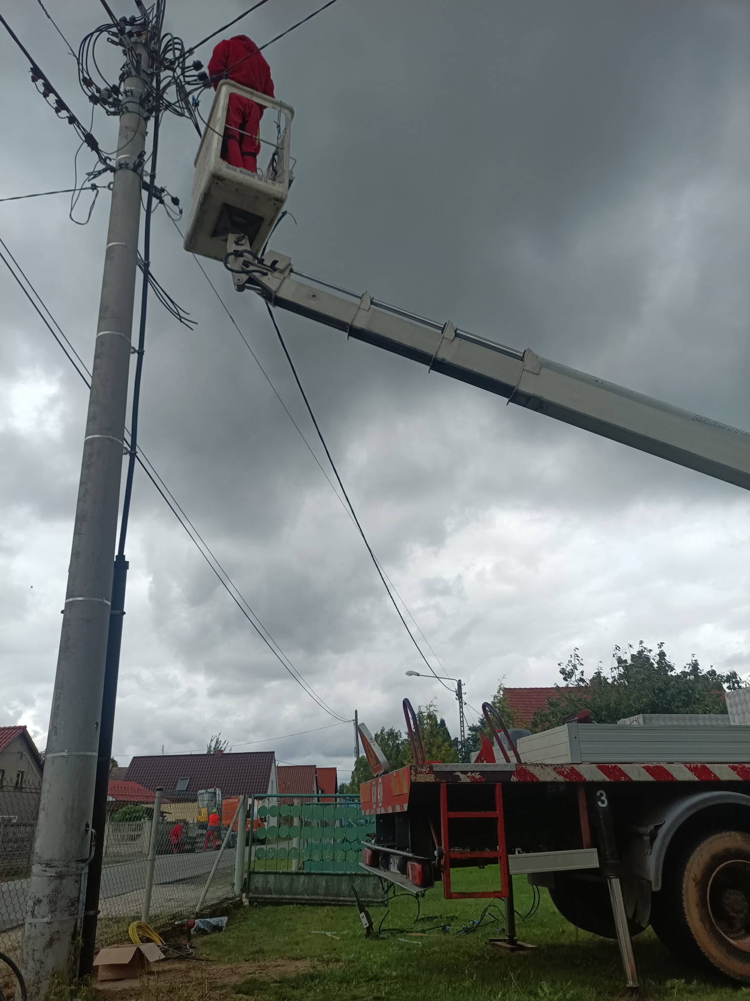 Pracownik w czerwonym kombinezonie w koszu podnośnika na tle pochmurnego nieba, wykonujący prace przy słupie energetycznym z gęstą siecią kabli.