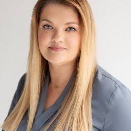 Centrum Hipnoterapii MyPRO Therapy - Portret kobiety o blond włosach i niebieskich oczach, ubrana w szarą bluzkę, z delikatnym uśmiechem na jasnym tle. Widoczny złoty naszyjnik.