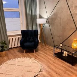 Centrum Hipnoterapii MyPRO Therapy - Stylowe wnętrze pokoju z ciemnym fotelem, lampą stojącą i okrągłym dywanem. Na ścianie dekoracyjny wzór z czarnych linii. Przytulna aranżacja z elementami dekoracyjnymi.