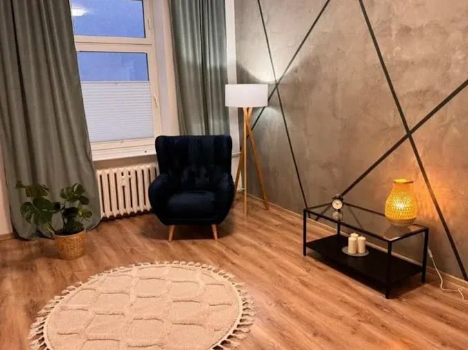 Stylowe wnętrze pokoju z ciemnym fotelem, lampą stojącą i okrągłym dywanem. Na ścianie dekoracyjny wzór z czarnych linii. Przytulna aranżacja z elementami dekoracyjnymi.