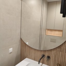 Pracownia meblarska Patryk suwała - Nowoczesna łazienka z owalnym lustrem, beżowe płytki na ścianach, biała umywalka i szafka. Minimalistyczny design z akcentami drewna i czarnym oświetleniem.