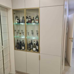 Pracownia meblarska Patryk suwała - Biała szafa wnękowa z przeszkloną witryną, podświetlona na zielono, eksponująca kolekcję alkoholi i kieliszków, z minimalistycznymi złotymi uchwytami.