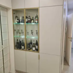 Biała szafa wnękowa z przeszkloną witryną, podświetlona na zielono, eksponująca kolekcję alkoholi i kieliszków, z minimalistycznymi złotymi uchwytami.