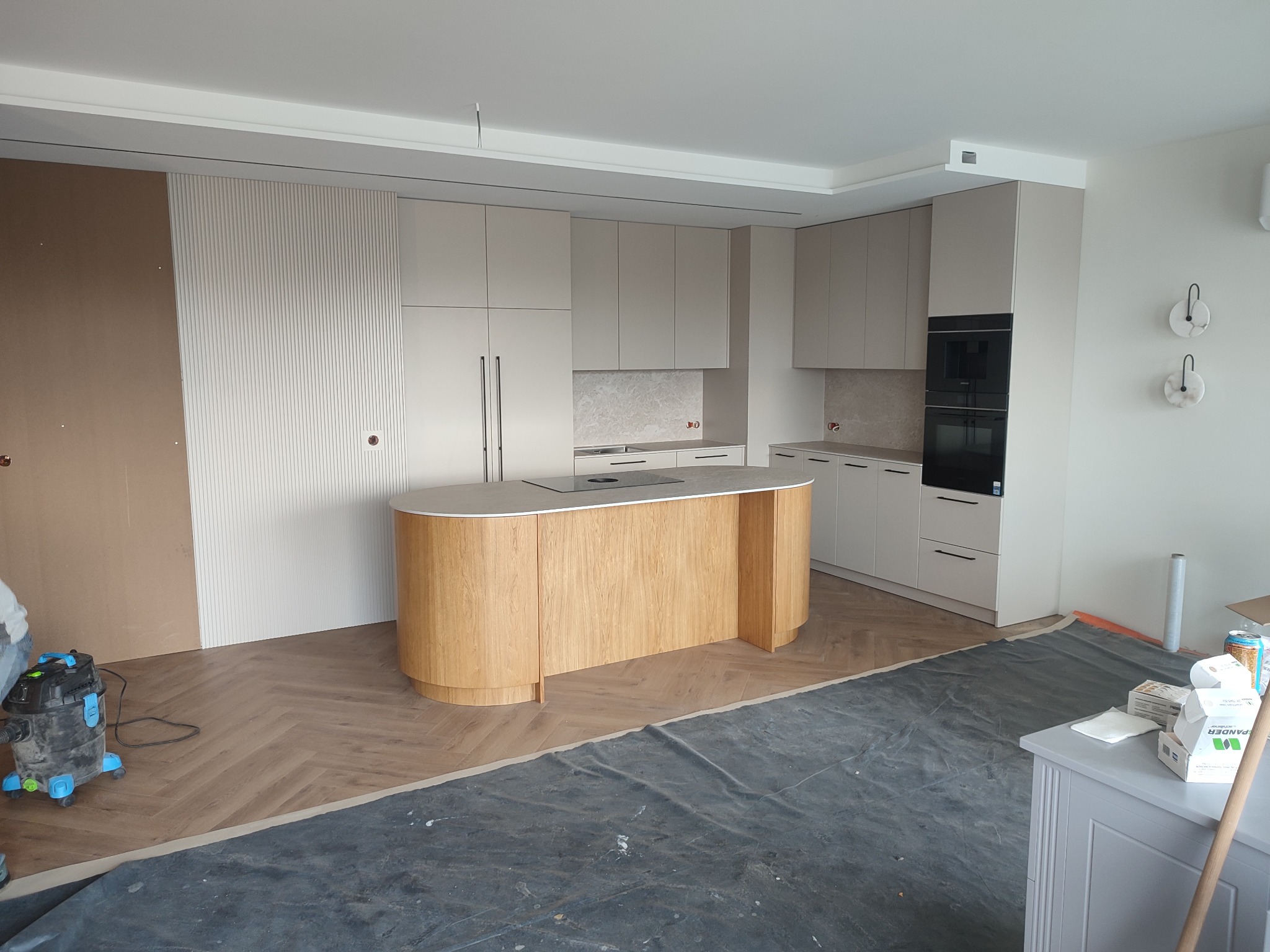 Jasna kuchnia z drewnianą wyspą i sprzętem AGD w zabudowie. Parkiet ułożony w jodełkę. Częściowo zabezpieczona folią podłoga sugeruje trwający montaż.