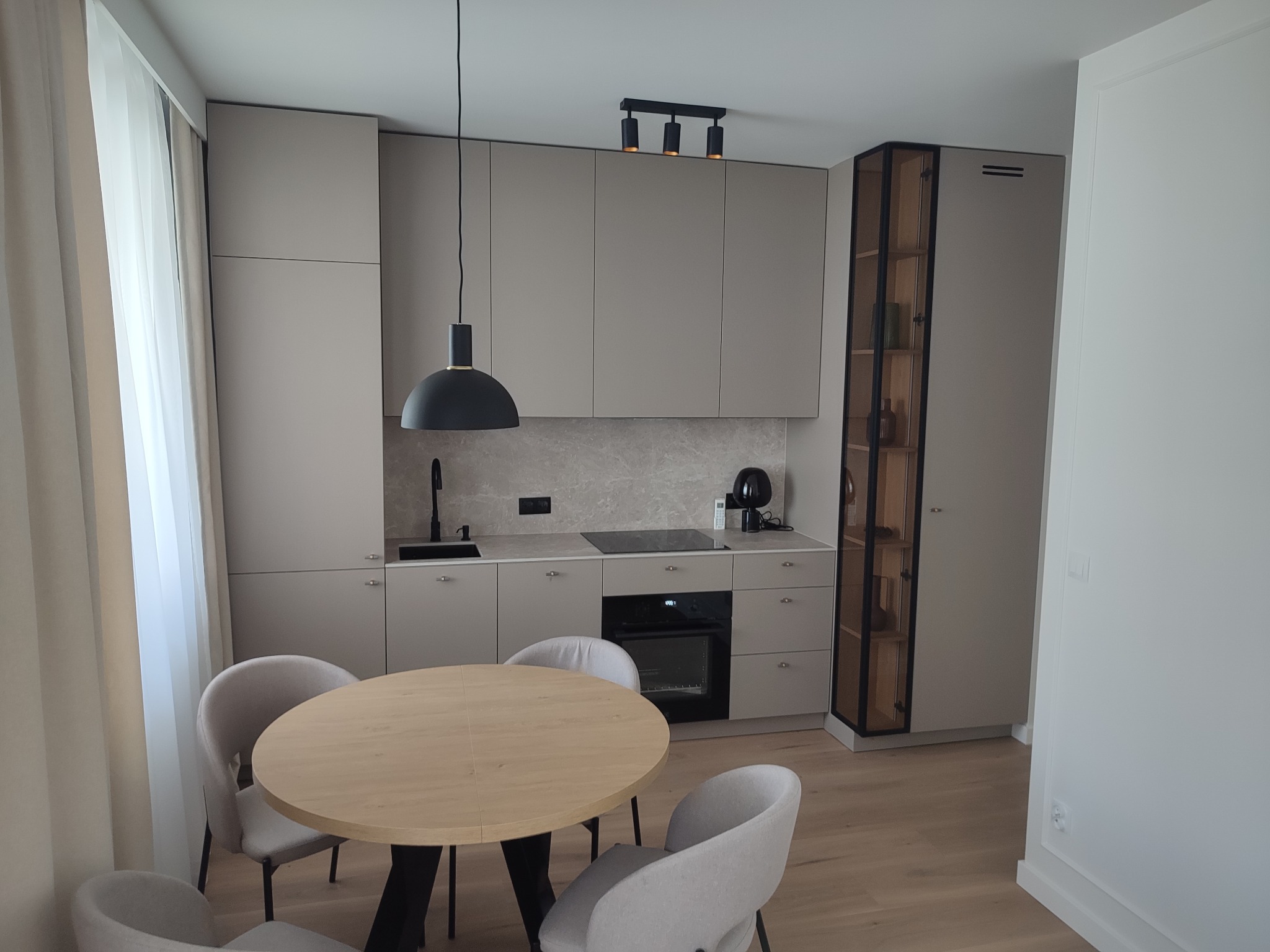Jasna kuchnia z okrągłym stołem i krzesłami. Zabudowa w odcieniach szarości z czarnymi akcentami oświetleniowymi. Na blacie widoczne akcesoria kuchenne.