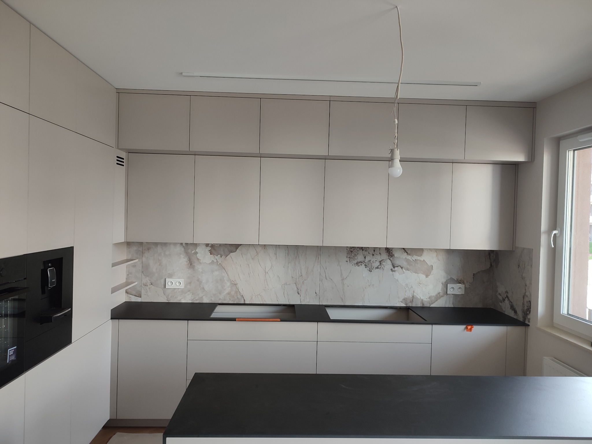 Jasna kuchnia z czarnym blatem i marmurowym panelem ściennym. Minimalistyczny design, wisząca żarówka bez klosza. Nowoczesne AGD w zabudowie.