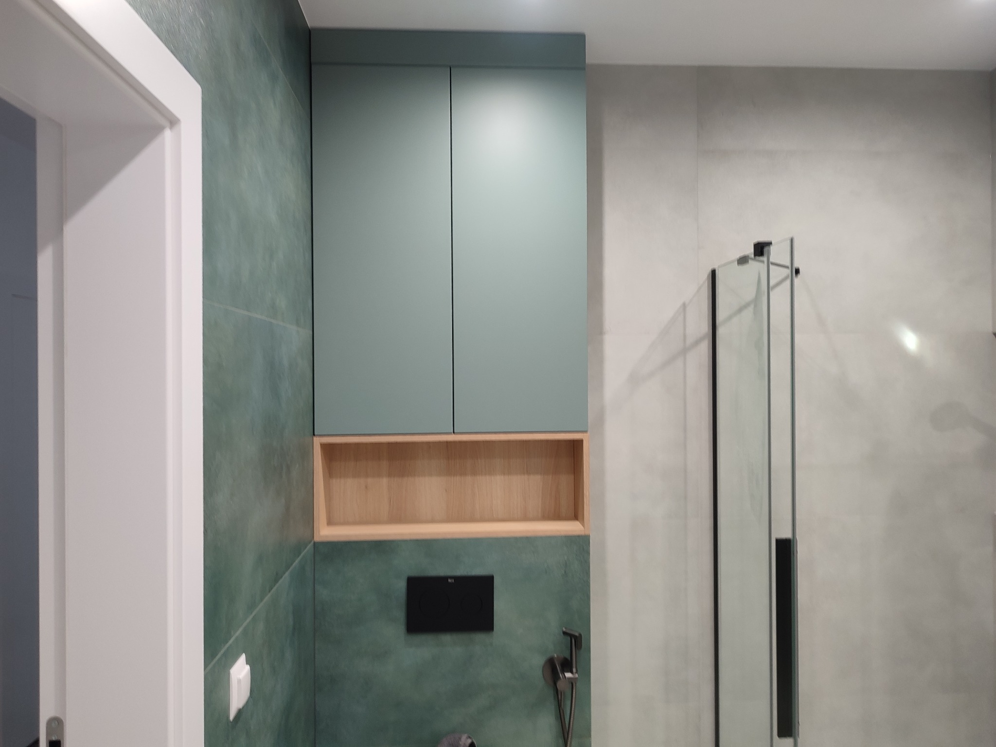 Nowoczesna szafka łazienkowa w odcieniach zieleni i drewna, z otwartą półką. Uwagę przyciągają płytki w kolorze butelkowej zieleni i minimalistyczny design.