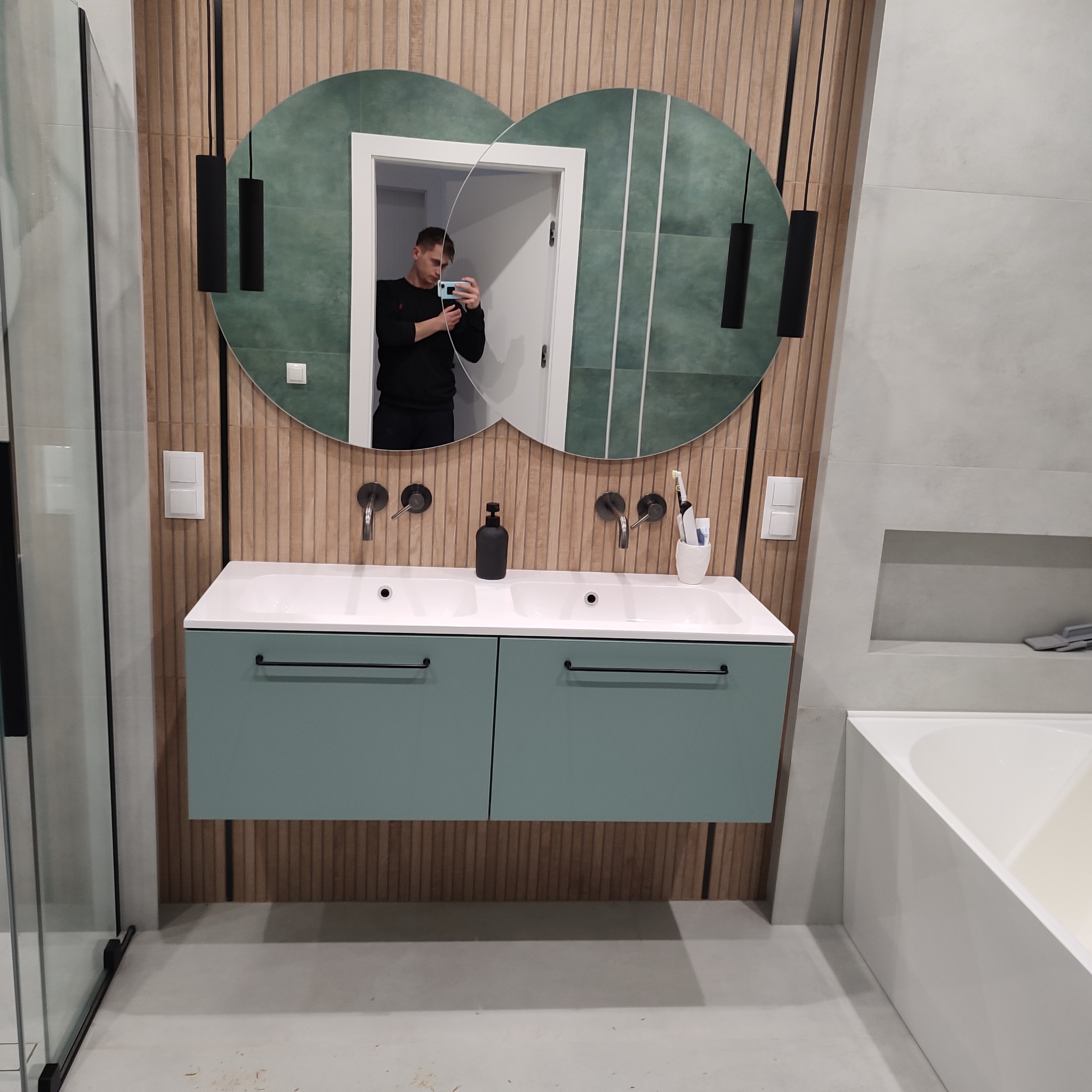Nowoczesna łazienka z podwójną umywalką, szafką w odcieniu zieleni i lustrem w kształcie dwóch kół. Widoczne drewniane panele na ścianie i czarne lampy wiszące.