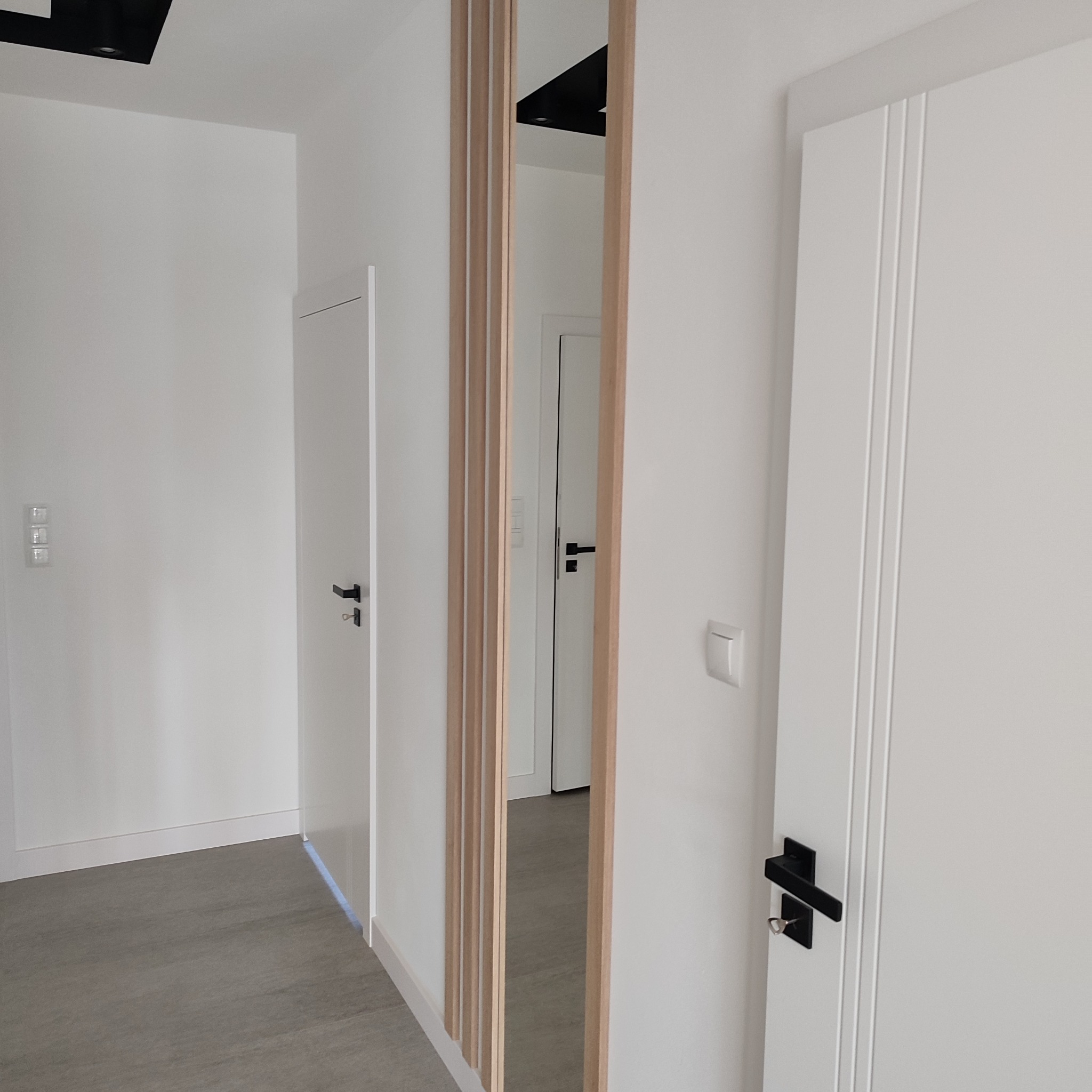 Nowoczesne wnętrze z białymi drzwiami z czarnymi klamkami i drewnianą ścianką działową z pionowych listew, widok na korytarz i kolejne drzwi.