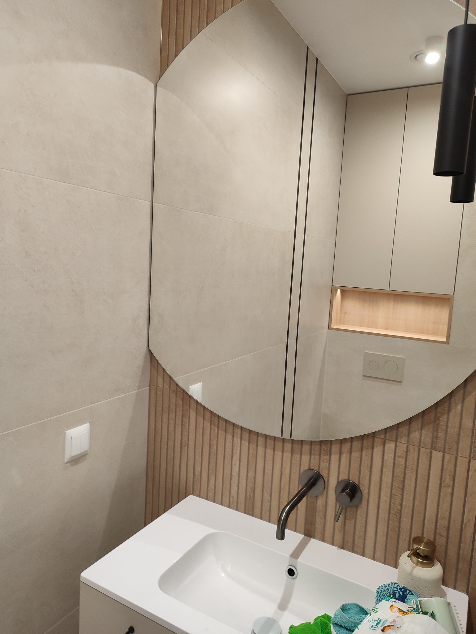 Nowoczesna łazienka z owalnym lustrem, beżowe płytki na ścianach, biała umywalka i szafka. Minimalistyczny design z akcentami drewna i czarnym oświetleniem.