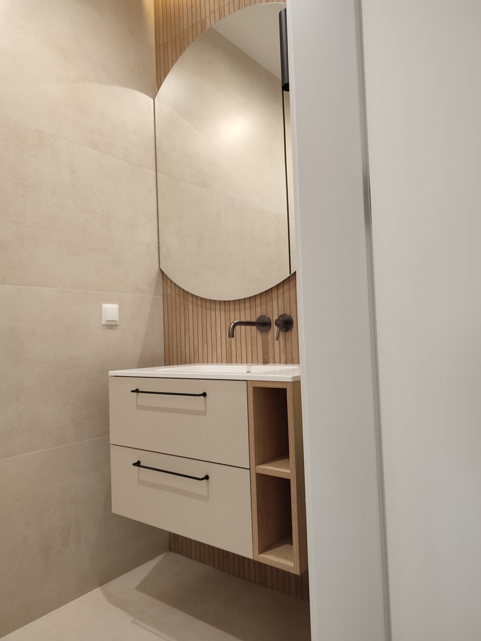 Nowoczesna łazienka: beżowa szafka z szufladami i półkami, okrągłe lustro, drewniane panele na ścianie. Minimalistyczny design, stonowana kolorystyka, czarne uchwyty i bateria.