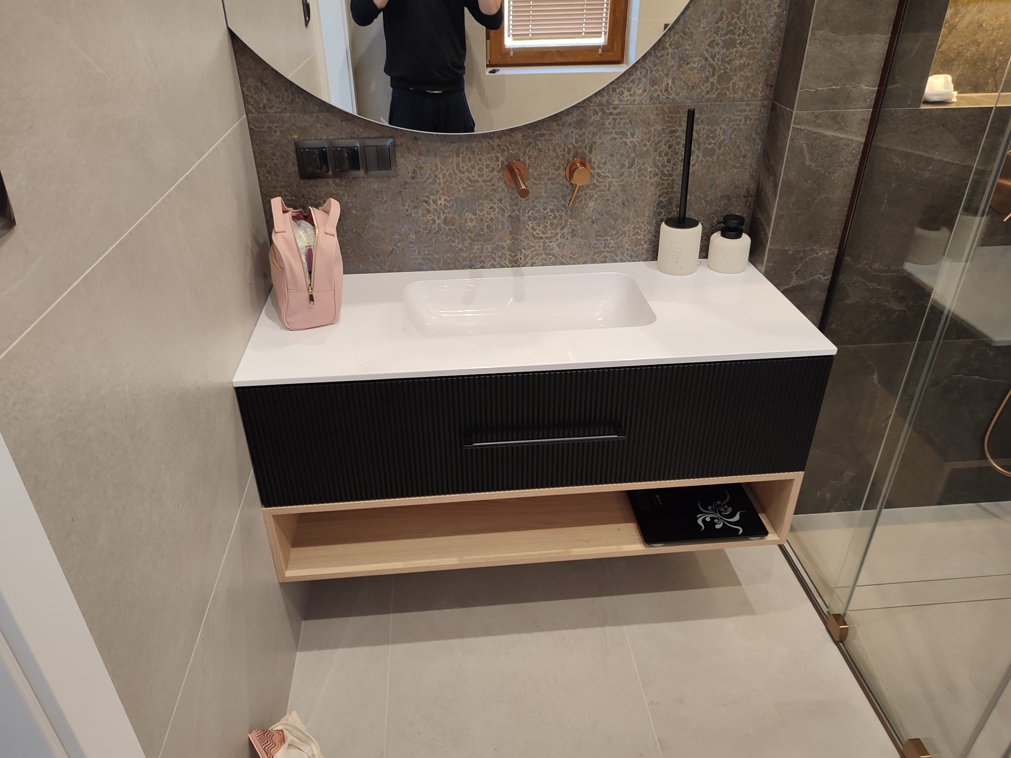 Nowoczesna szafka łazienkowa z czarnym frontem ryflowanym i jasnym blatem, z owalnym lustrem i miedzianą armaturą na tle mozaiki w odcieniach szarości.