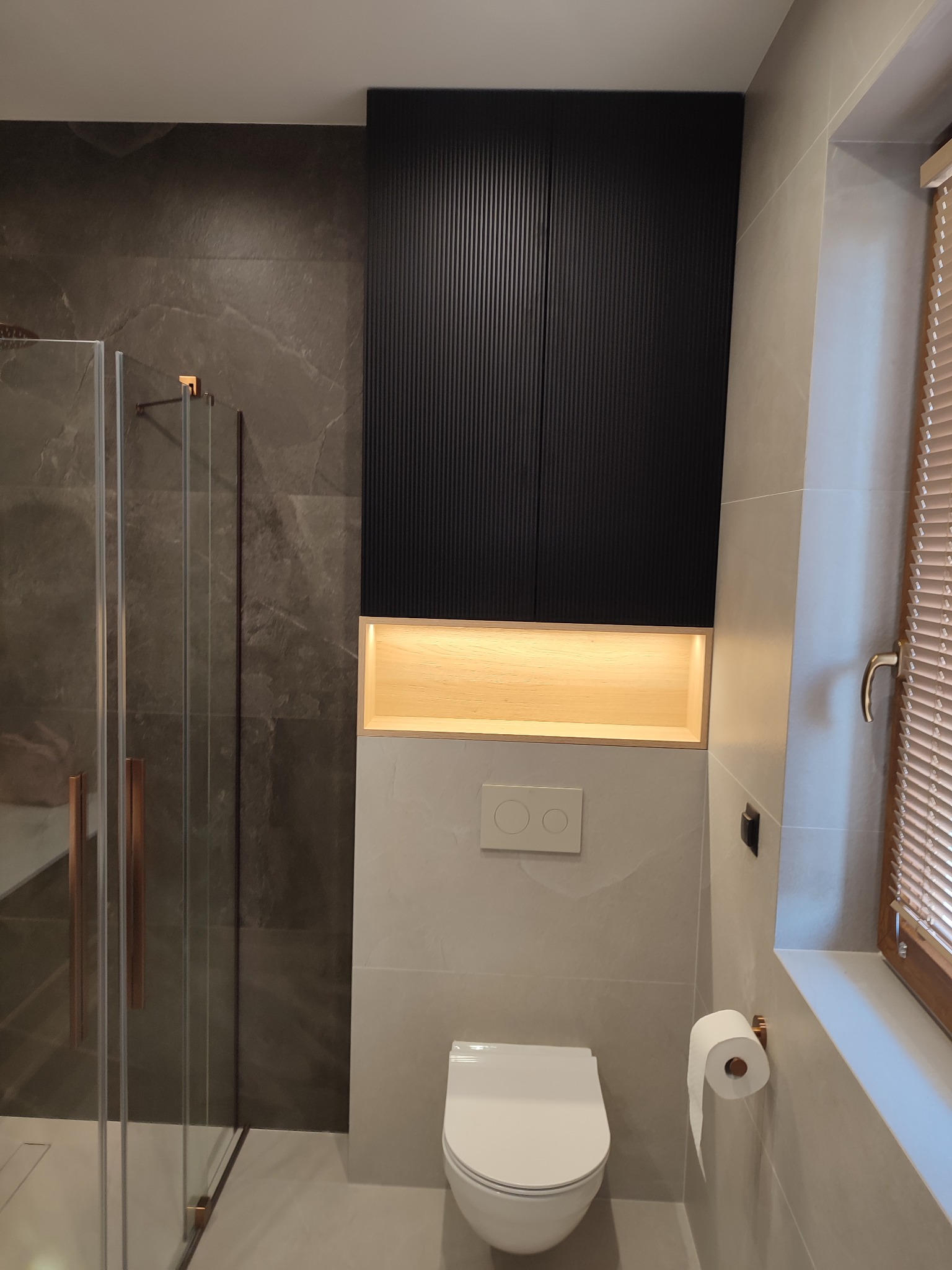 Nowoczesna łazienka: czarna szafka z oświetleniem nad toaletą, szklana kabina prysznicowa z miedzianymi uchwytami, rolety drewniane w oknie.