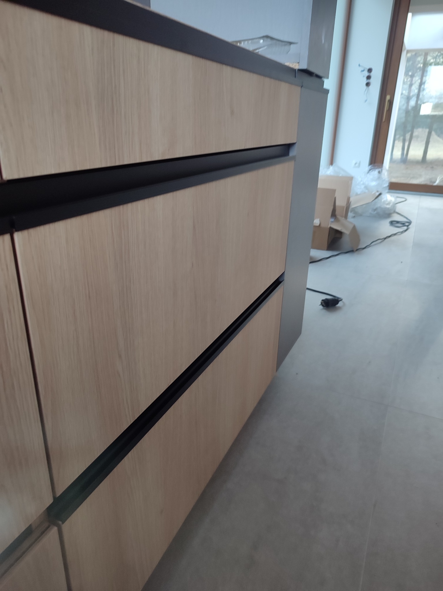 Front szuflad kuchennych z jasnego drewna z czarnymi uchwytami, widoczne z boku, z oknem i kartonami w tle, co sugeruje świeży montaż.