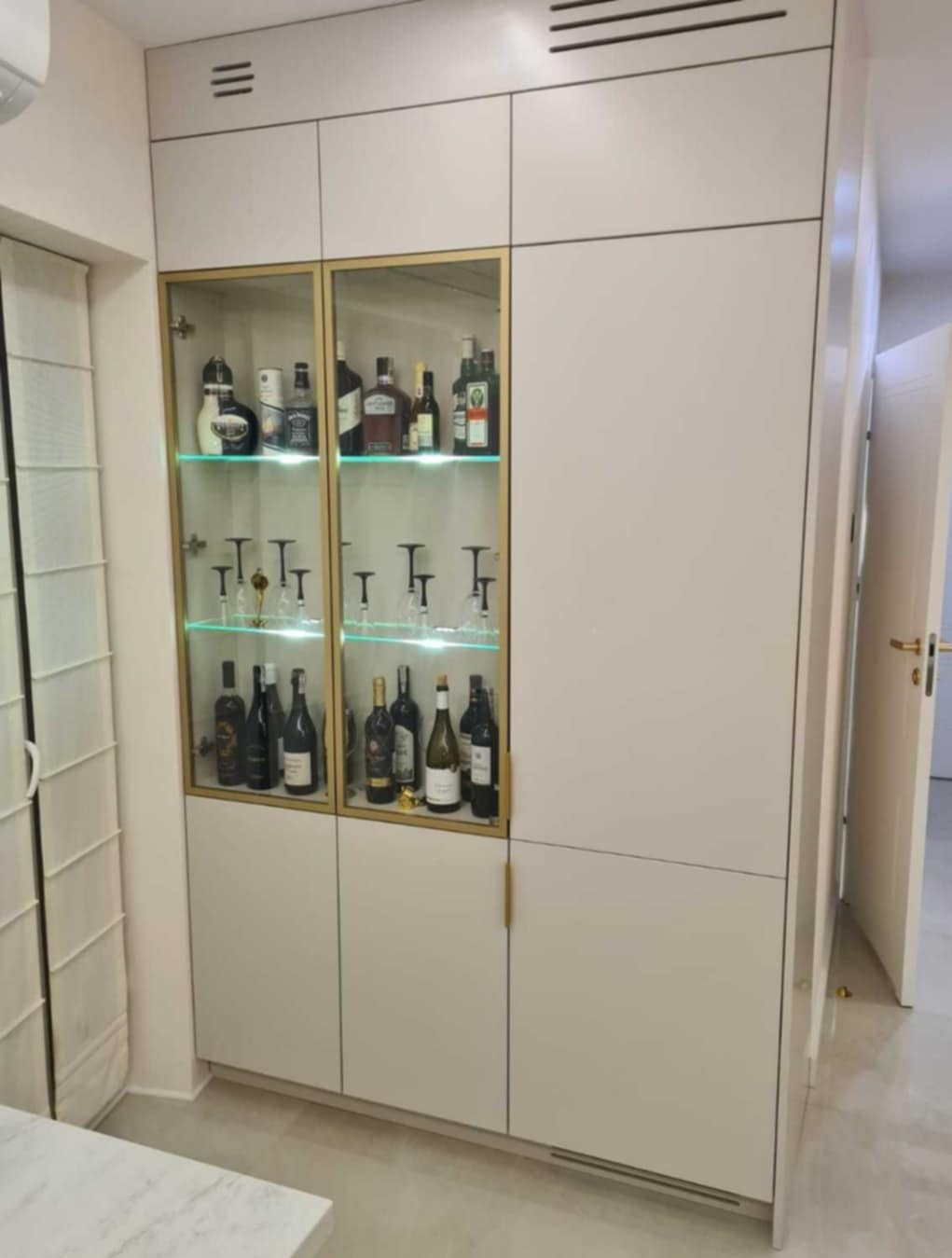 Biała szafa wnękowa z przeszkloną witryną, podświetlona na zielono, eksponująca kolekcję alkoholi i kieliszków, z minimalistycznymi złotymi uchwytami.