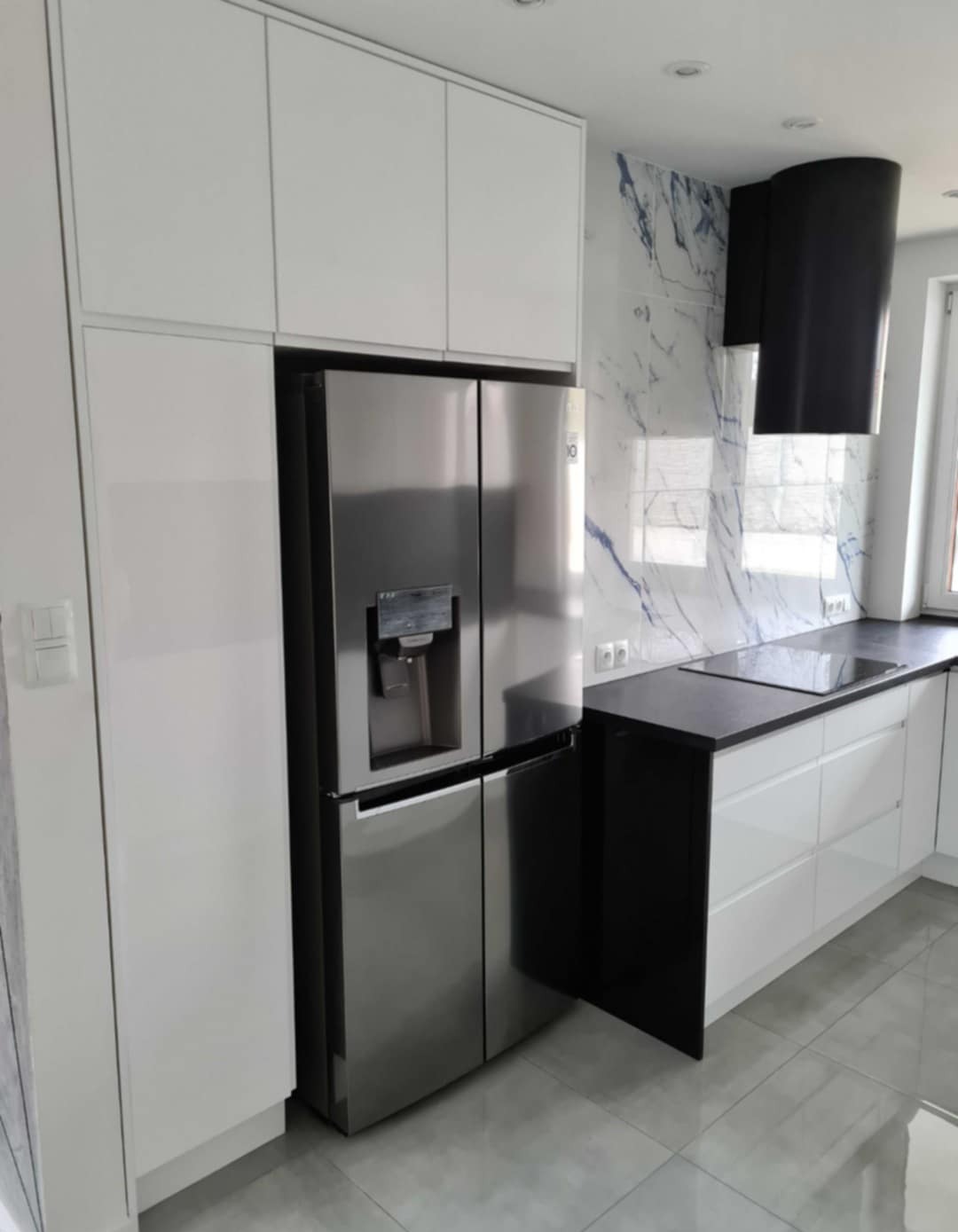 Nowoczesna kuchnia na wymiar: białe szafki zintegrowane z lodówką side-by-side, czarny blat i okap na tle marmurowej ściany z niebieskimi żyłami.