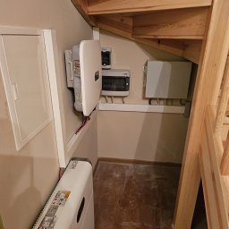 OZEHome Mykhailo Volkov - Instalacja magazynu energii pod drewnianymi schodami: biała obudowa, skrzynki elektryczne, kable. Nietypowe umiejscowienie, widoczne elementy instalacji.