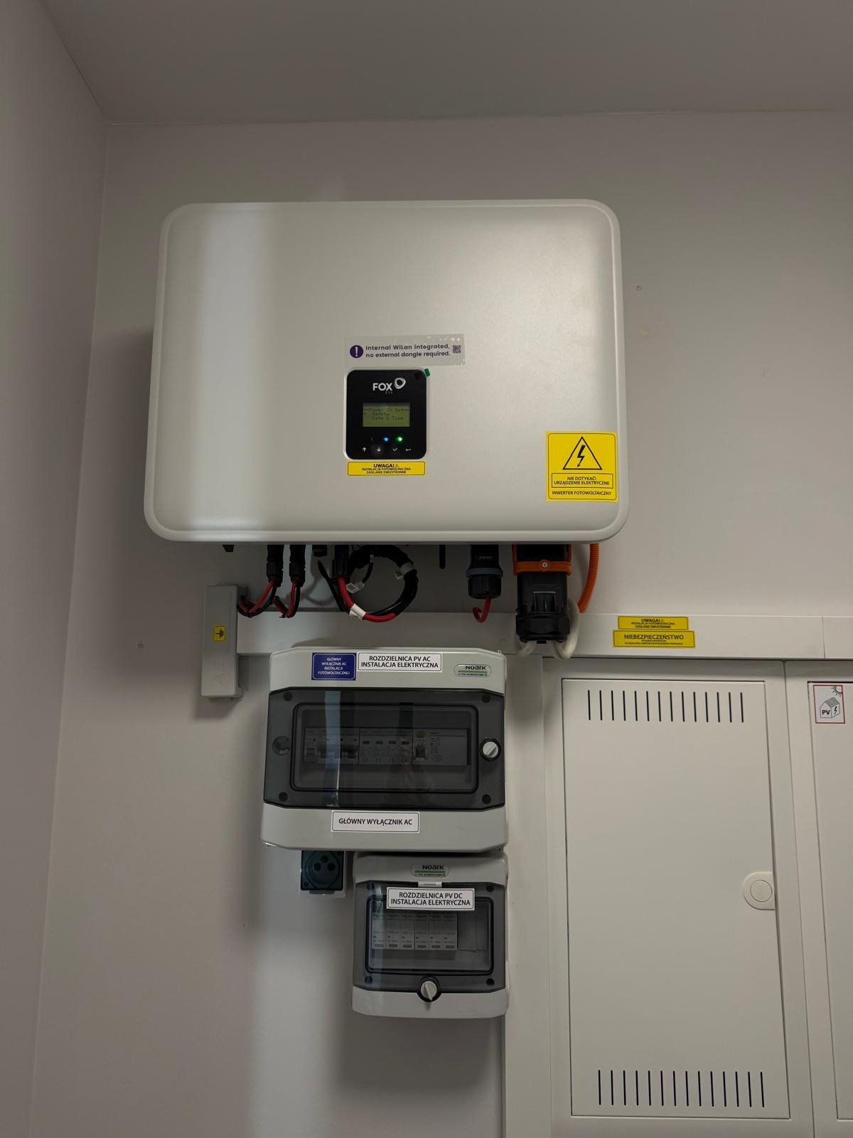 Zamontowany inwerter fotowoltaiczny Fox z rozdzielnicami AC/DC na ścianie. Widoczne okablowanie i naklejki ostrzegawcze. Instalacja wewnątrz budynku.