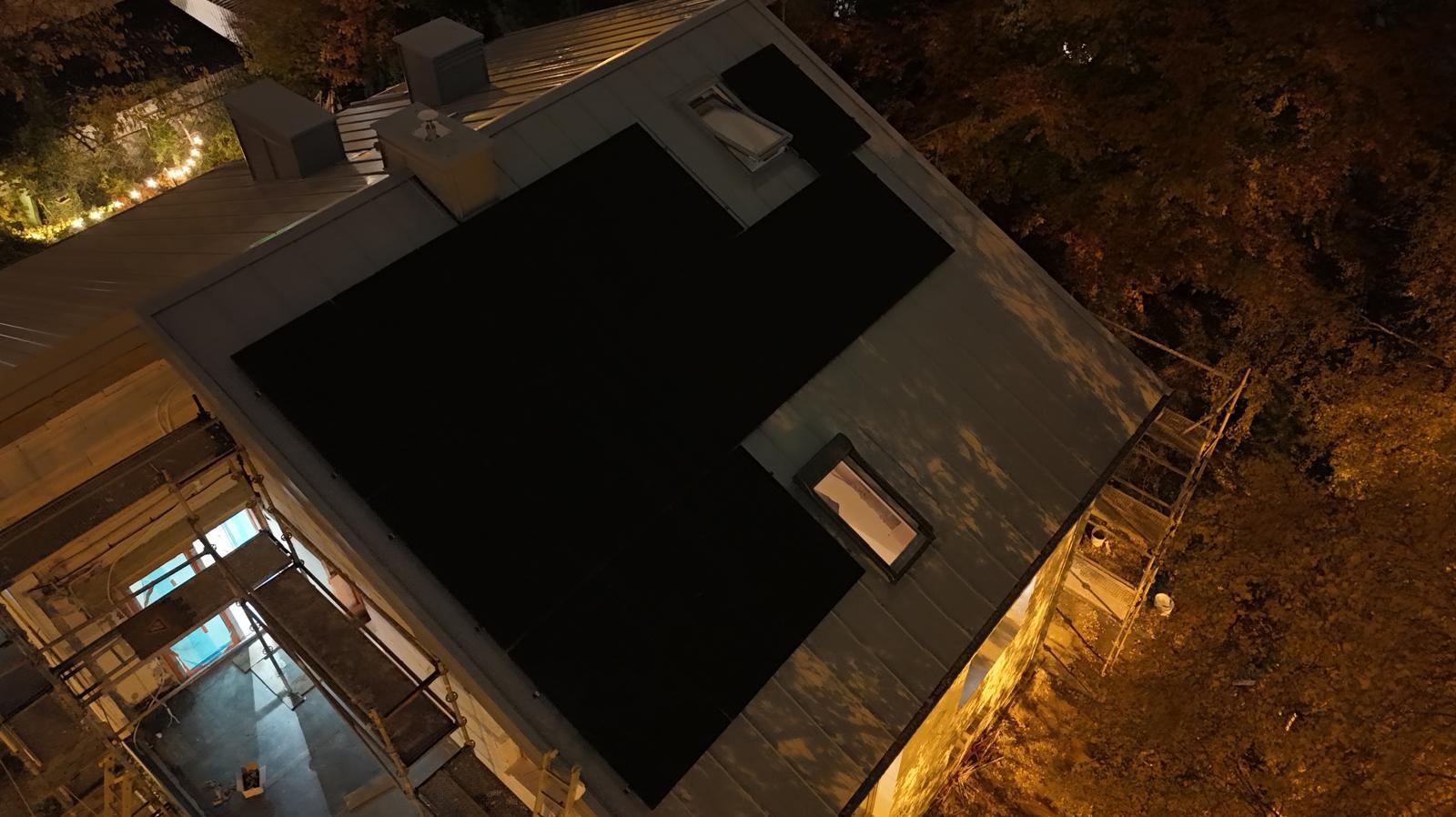 Nocny widok z góry na dach z panelami słonecznymi i oknami dachowymi, częściowo zasłonięty rusztowaniem. Oświetlenie nocne, drzewa w tle.