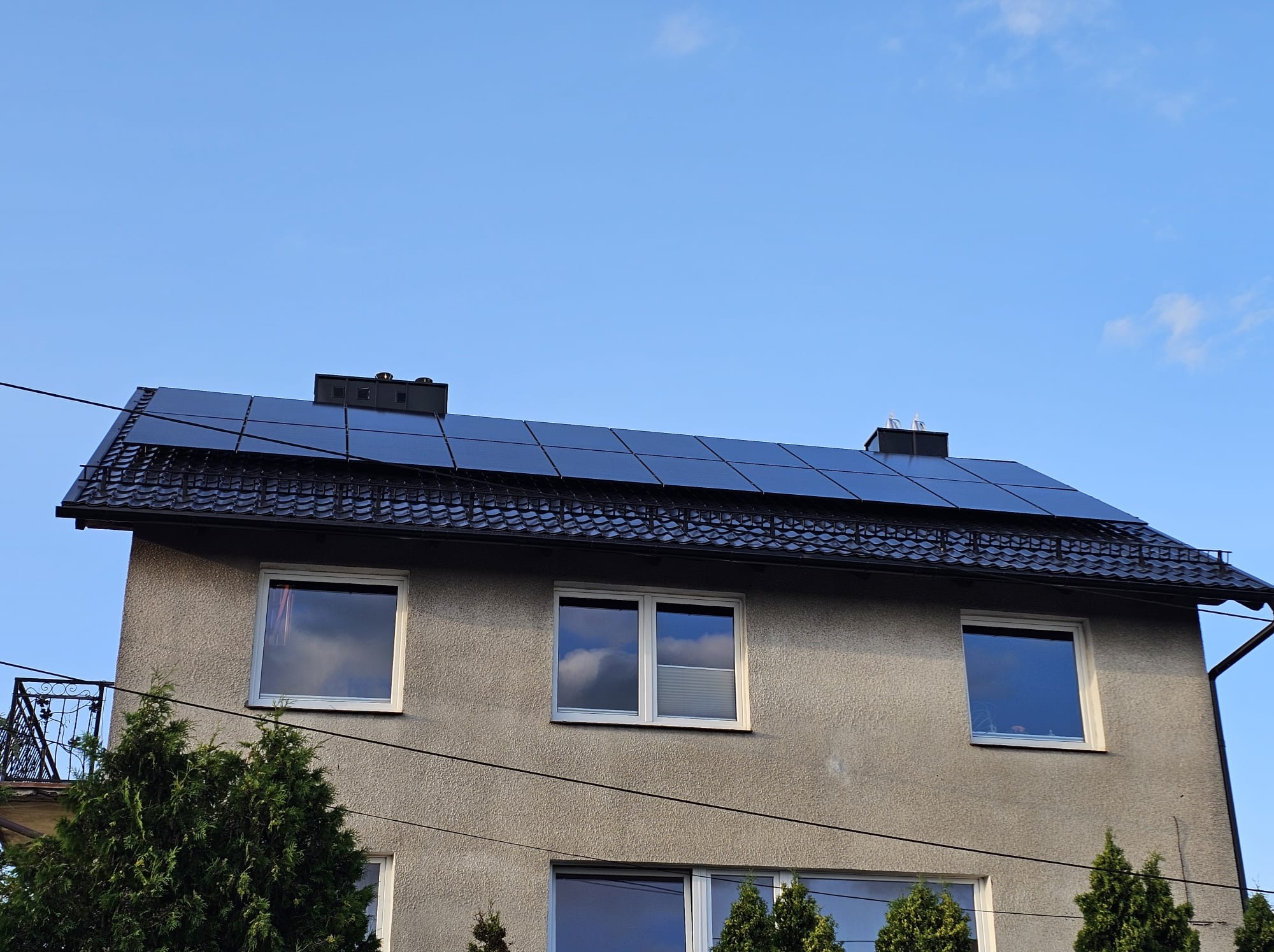 Dwupiętrowy dom z ciemnymi panelami fotowoltaicznymi na dachu, widocznymi na tle błękitnego nieba. Elewacja szara, okna z odbiciem nieba.