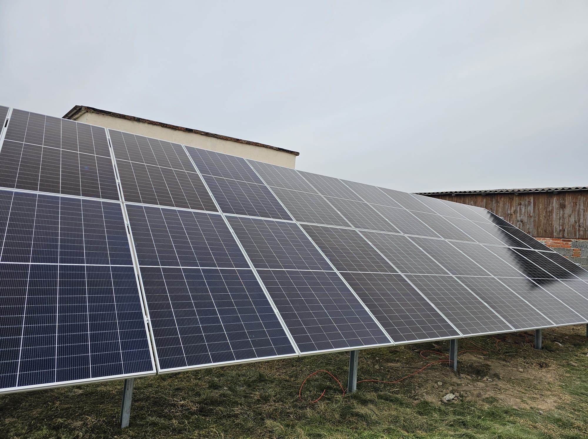 Instalacja fotowoltaiczna na gruncie, widok z boku. Panele zamontowane na metalowych słupkach, w tle budynek z cegły i drewniana konstrukcja. Pochmurne niebo.