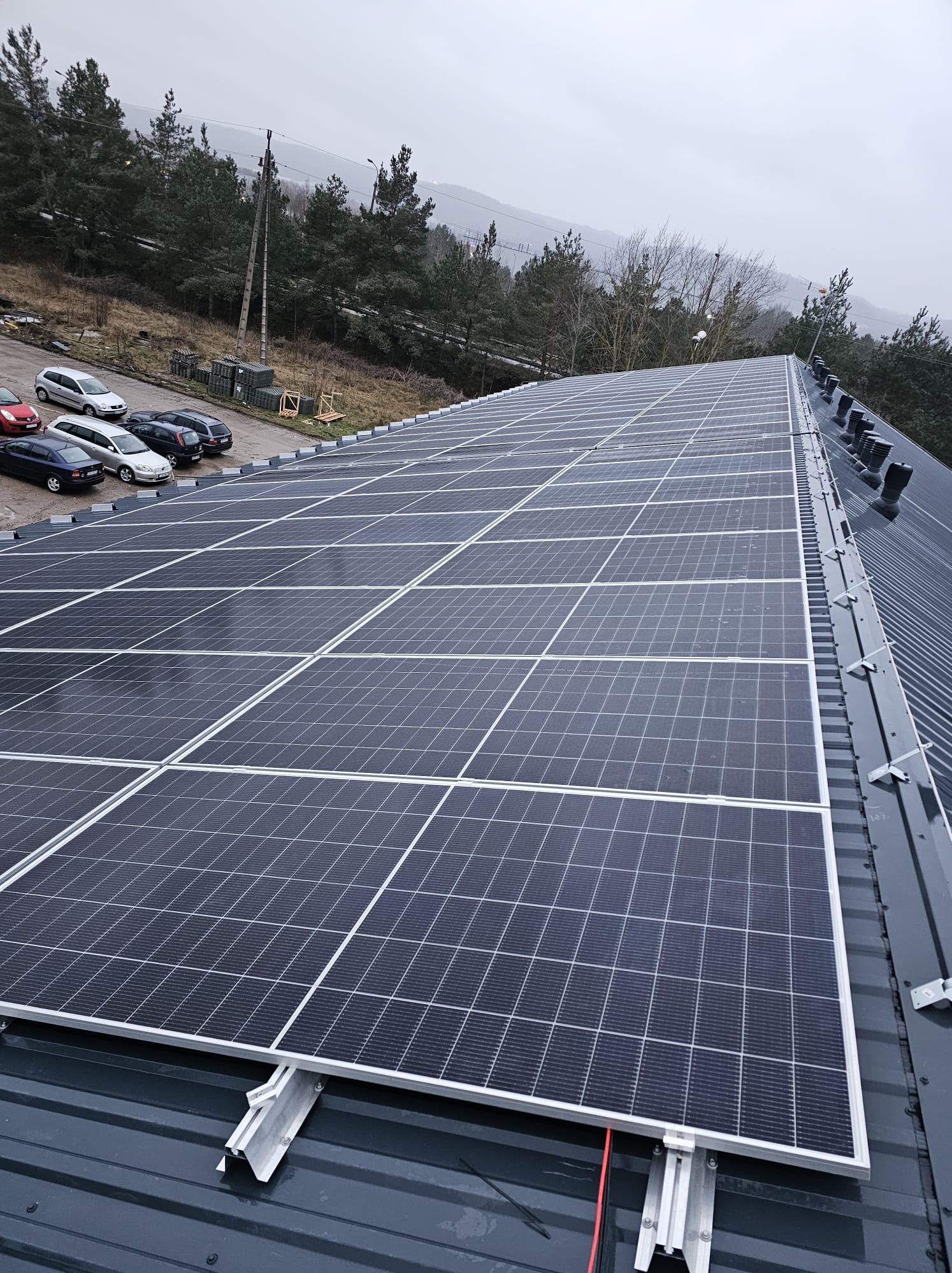 Panel słoneczny na dachu budynku przemysłowego, widok z góry, w tle zaparkowane samochody i drzewa. Montaż paneli fotowoltaicznych na dachu z blachy trapezowej.