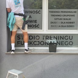 Dziemba Ci Umyje - Mężczyzna w koszulce z logo firmy czyści okno z napisem 'Studio Treningu Funkcjonalnego'. Widoczna drabina i wiadro. Ujęcie z zewnątrz budynku.