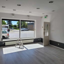 Dziemba Ci Umyje - Jasne wnętrze po sprzątaniu: białe ściany, szare panele, drabina pod oknem, widok na ulicę. Czysta przestrzeń gotowa do użytku, z wyraźnym światłem słonecznym.