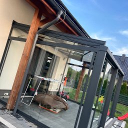 Dziemba Ci Umyje - Szklana zabudowa tarasu z szarymi ramami, widok z zewnątrz. Wewnątrz suszarka na pranie i inne przedmioty. Kostka brukowa na tarasie. Błękitne niebo w tle.