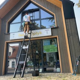 Dziemba Ci Umyje - Mężczyzna na drabinie myje okna witryny salonu optycznego z logotypami luksusowych marek. Budynek o nowoczesnej architekturze z elementami drewna.
