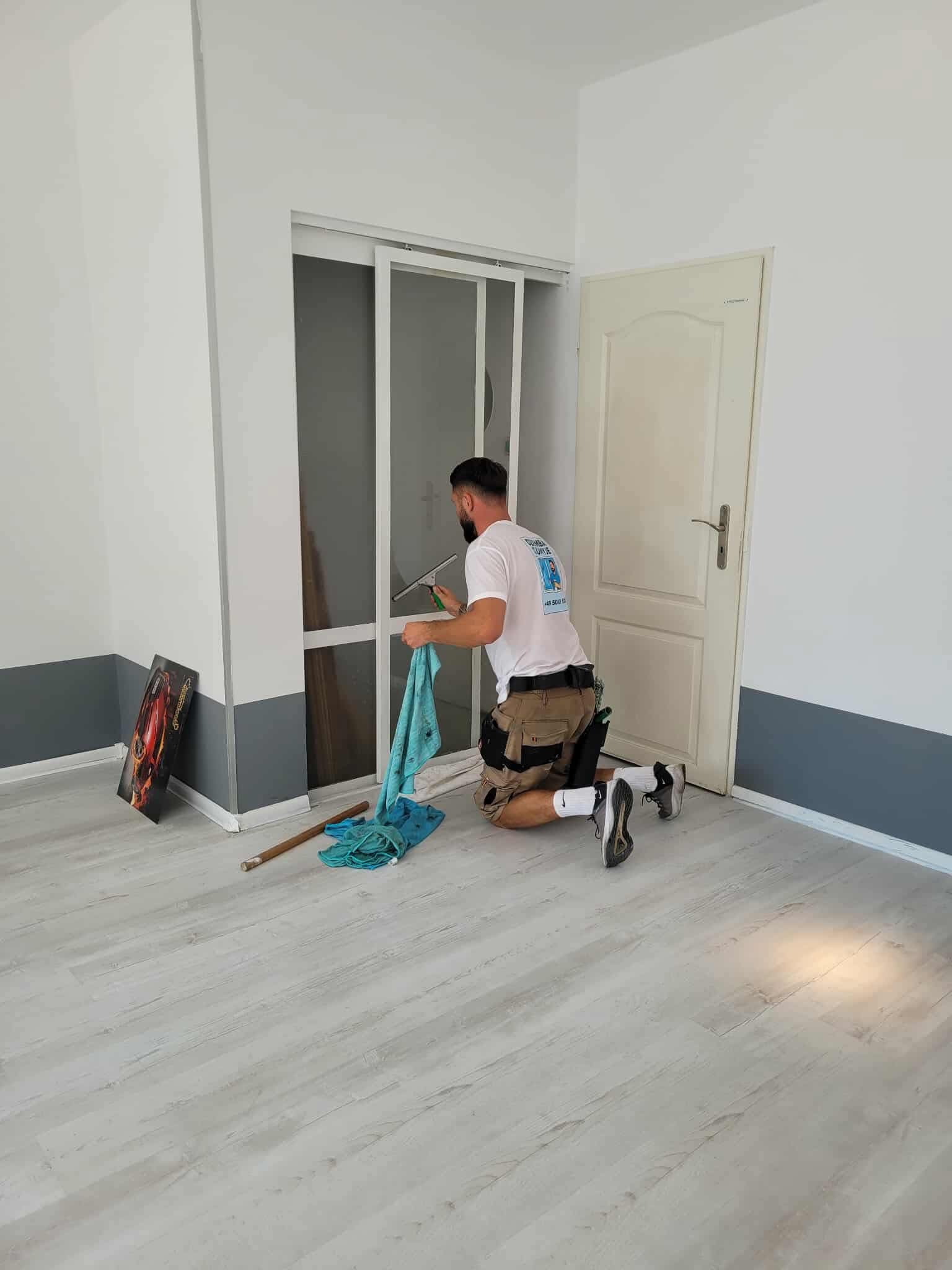 Mężczyzna w koszulce z logo firmy, klęczy i myje okno w mieszkaniu. Na podłodze leżą narzędzia i ściereczka. Widoczny obraz oparty o ścianę.