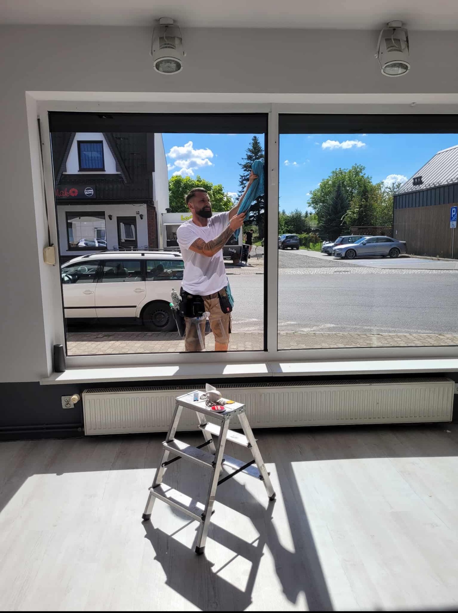 Mężczyzna z tatuażami myje okno w budynku, używając niebieskiej ściereczki. Na podłodze stoi drabina z narzędziami. Widok na ulicę z samochodami.