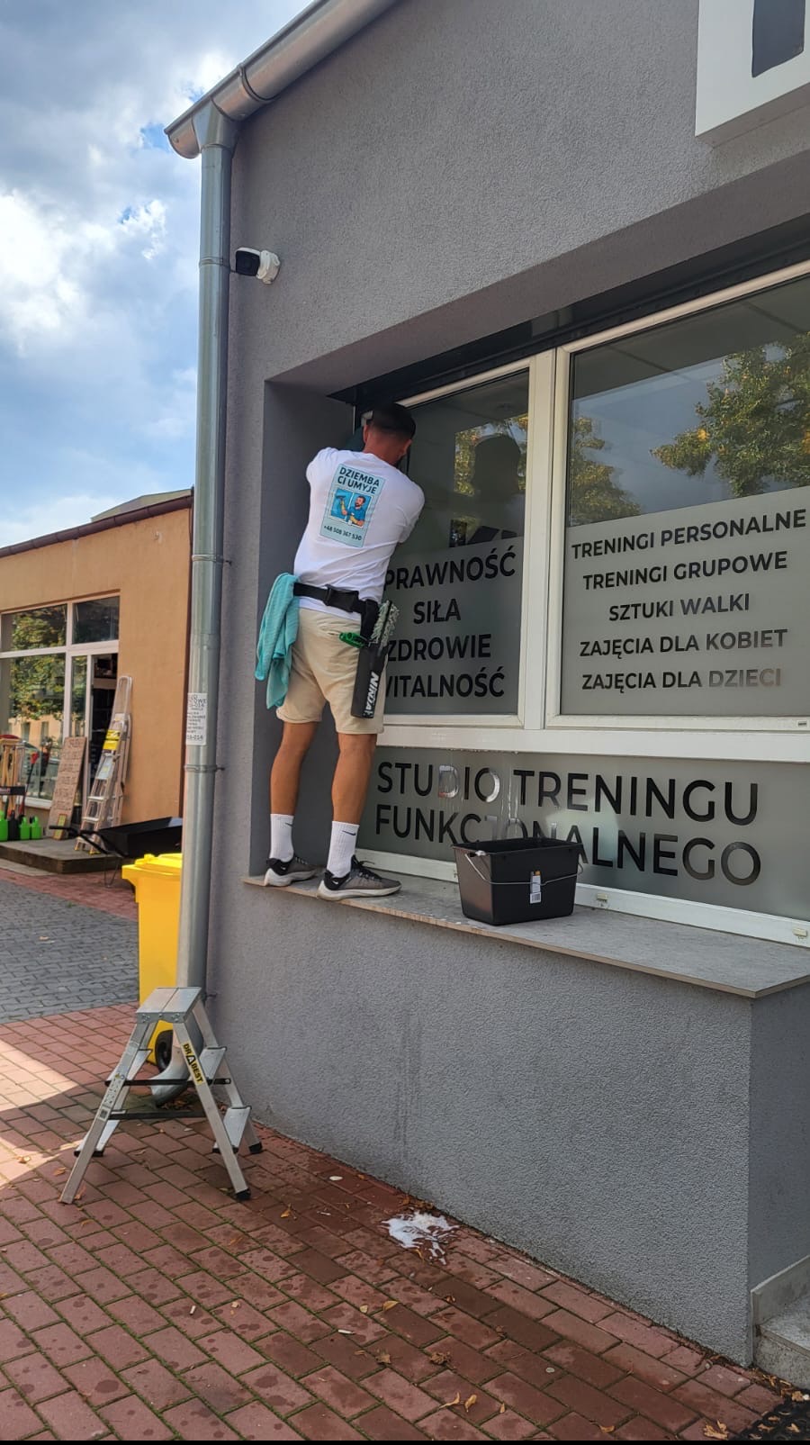 Mężczyzna w koszulce z logo firmy myje okno lokalu z napisem 'Studio Treningu Funkcjonalnego'. Na parapecie stoi wiadro, obok drabinka.