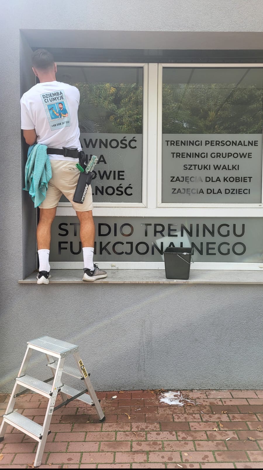 Mężczyzna w koszulce z logo firmy czyści okno z napisem 'Studio Treningu Funkcjonalnego'. Widoczna drabina i wiadro. Ujęcie z zewnątrz budynku.