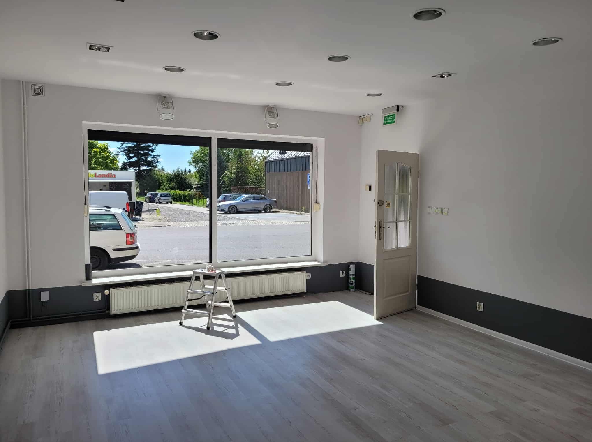 Jasne wnętrze po sprzątaniu: białe ściany, szare panele, drabina pod oknem, widok na ulicę. Czysta przestrzeń gotowa do użytku, z wyraźnym światłem słonecznym.
