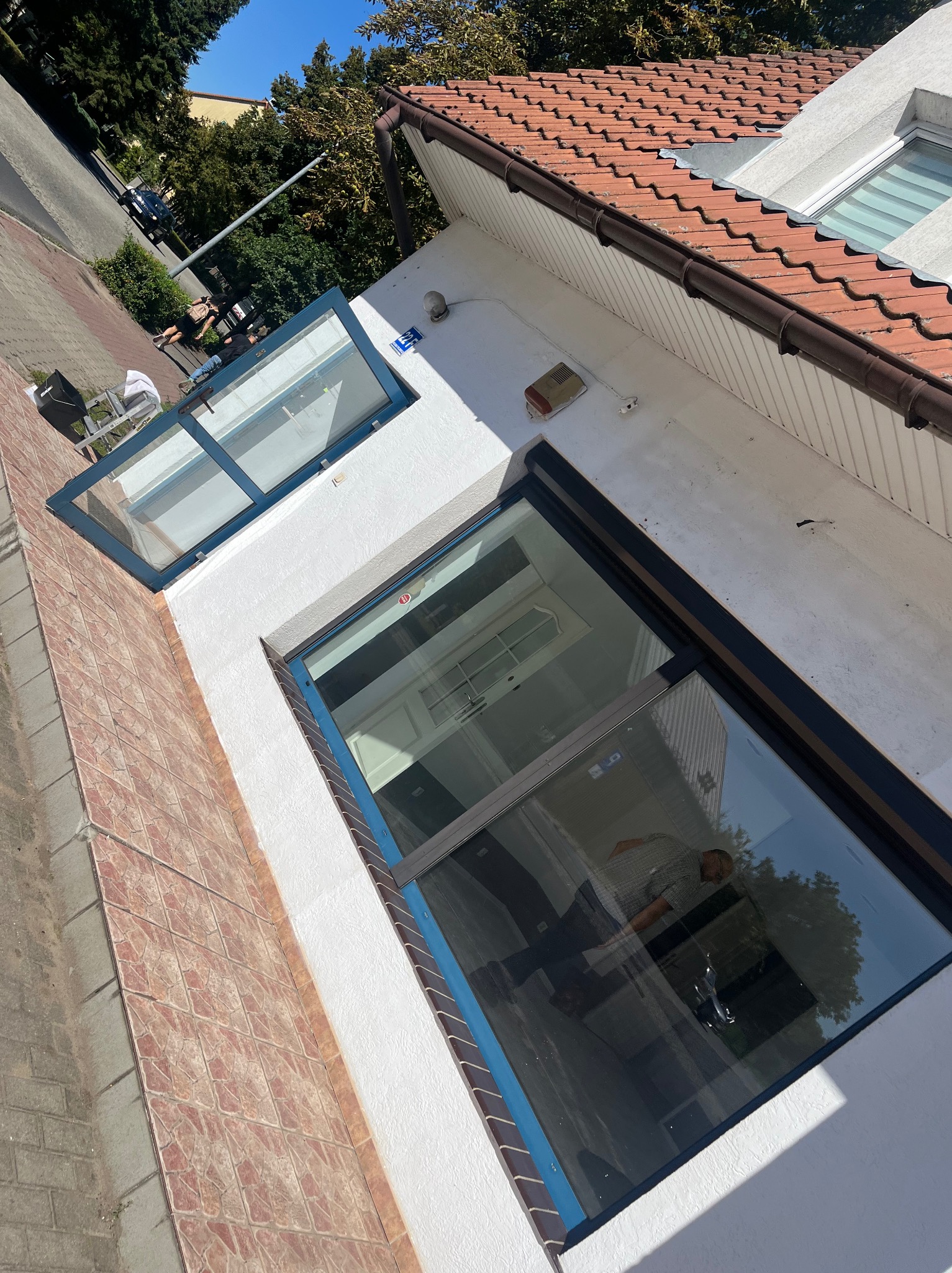 Ukośne ujęcie dwukondygnacyjnego budynku z oknami; w dolnym odbija się osoba, górne okno dachowe z roletami. Czerwona dachówka i biała elewacja.