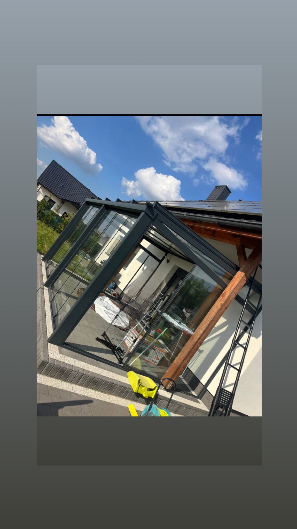 Szklana weranda z ciemną ramą, widok z zewnątrz. Na podłodze leżą akcesoria do sprzątania, a wewnątrz widoczna drabina. W tle dom i błękitne niebo.